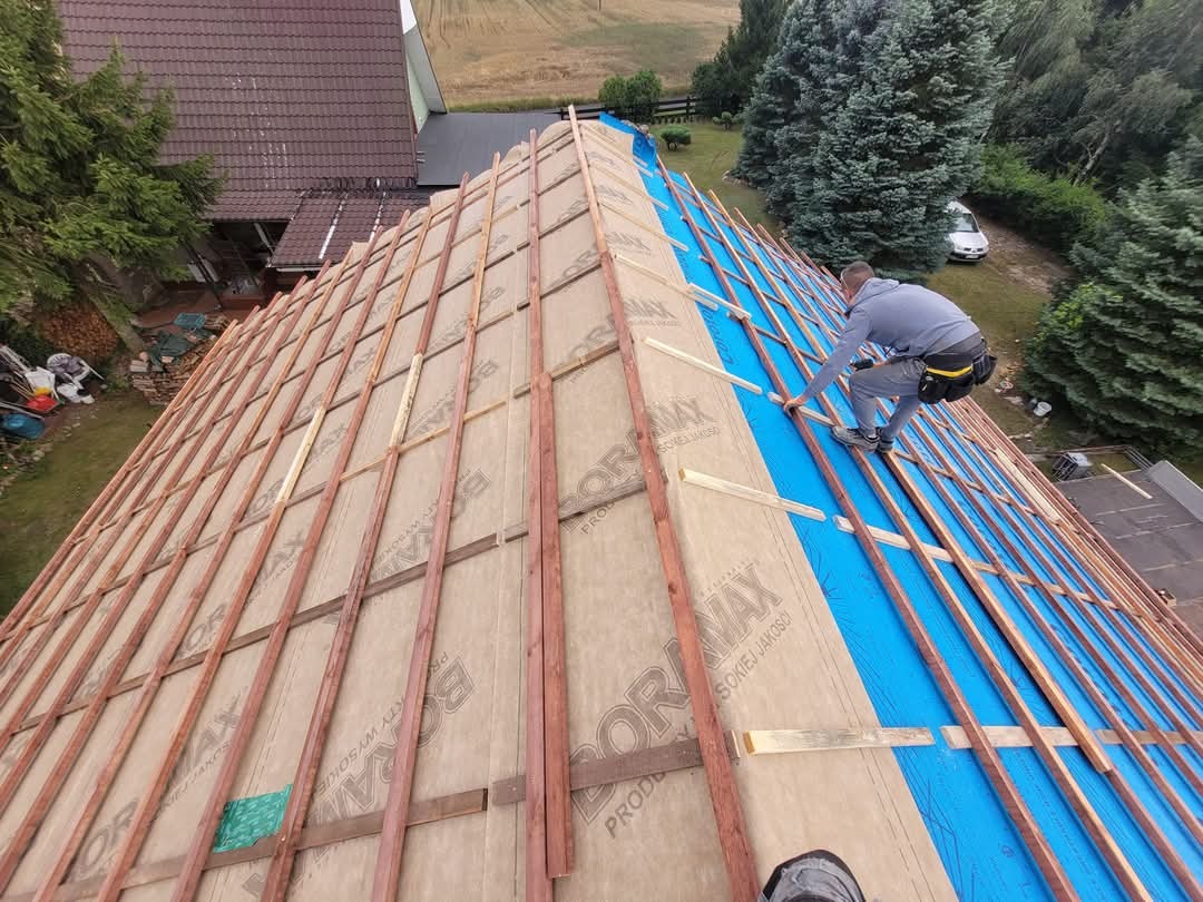Dekarz montuje kontrłaty na membranie dachowej, widok z góry na rozległy dach w trakcie prac, w tle dom i zieleń.