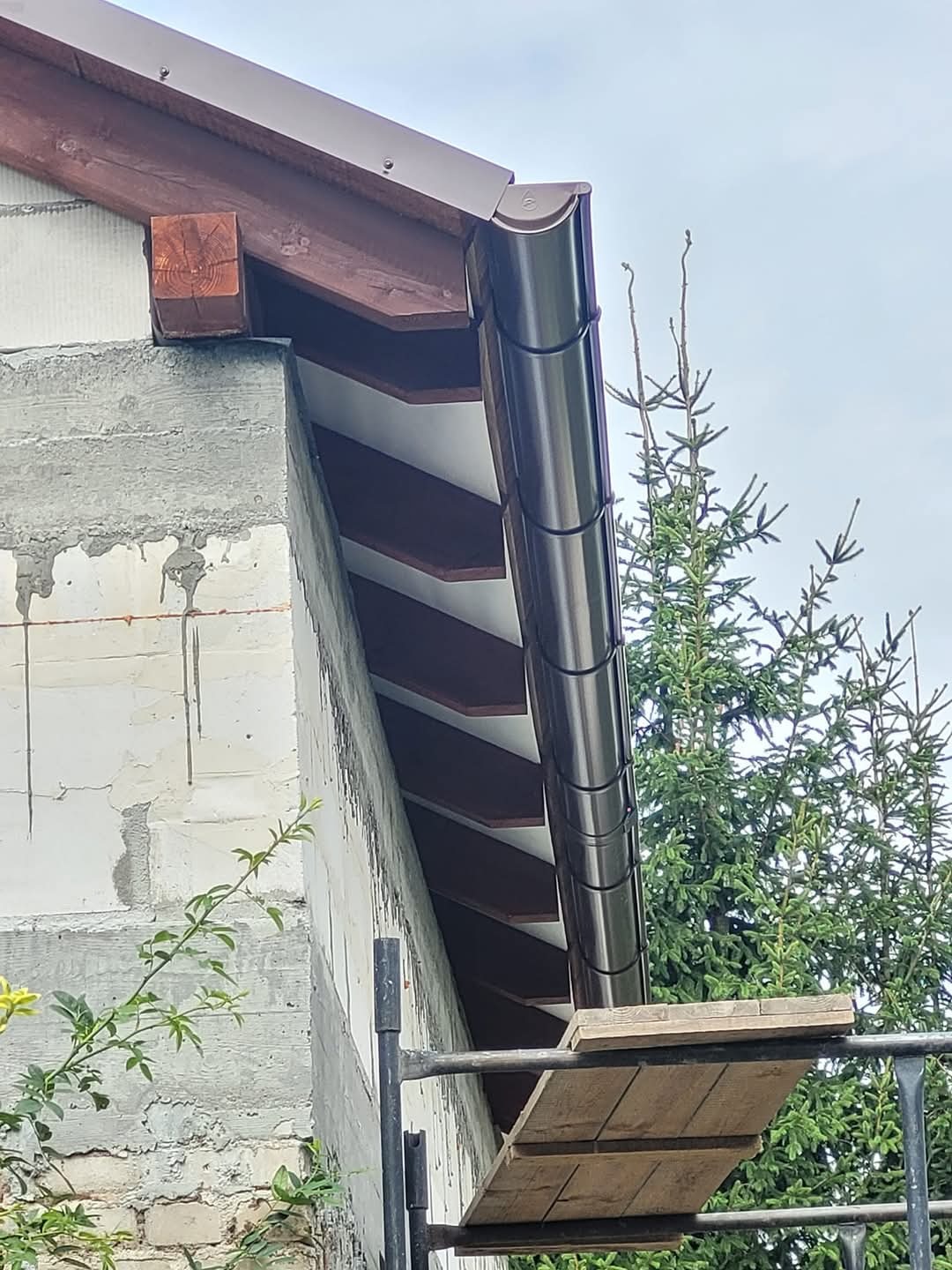 Zamontowana, brązowa rynna dachowa z bliska, widoczna ze rusztowania na tle ściany z betonu komórkowego i iglastego drzewa.
