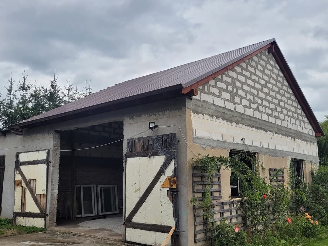 Budynek gospodarczy w Zakrzewie z nowym, brązowym dachem z blachy trapezowej i murem z betonu komórkowego w trakcie budowy, z otwartymi wrotami i oknami z kratami.