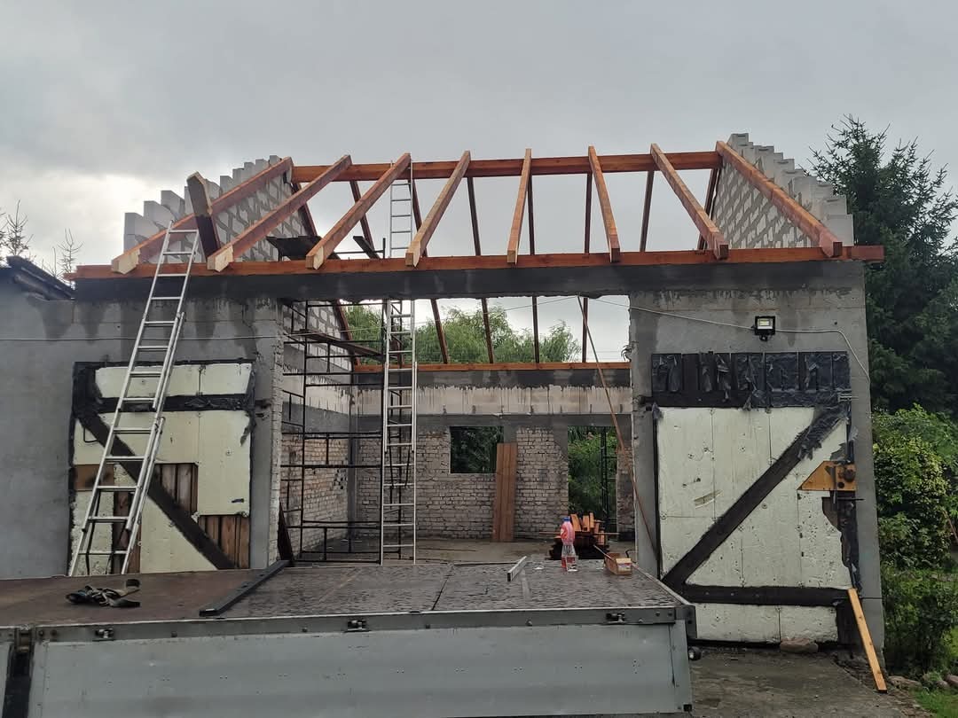 Budynek w trakcie budowy dachu, widoczne krokwie i pustaki, drabiny przystawione do ściany, na pierwszym planie platforma samochodu dostawczego.