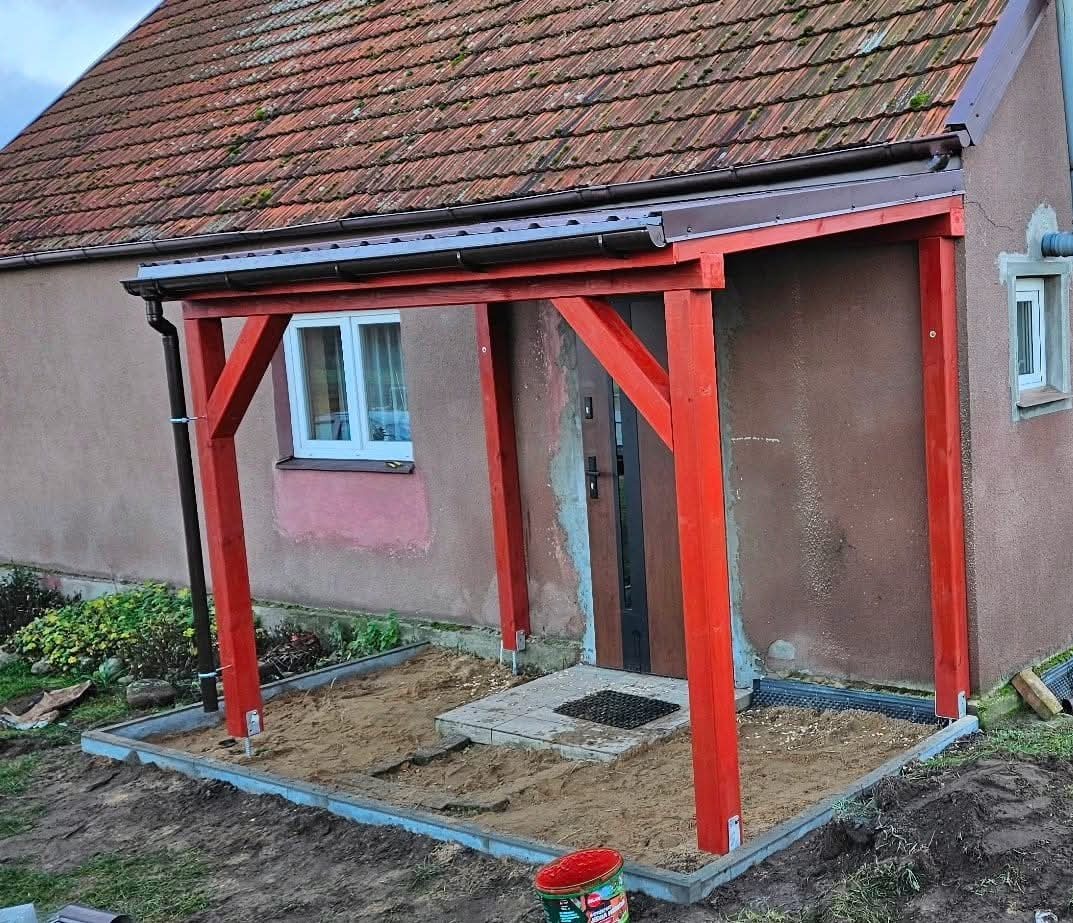 Czerwona drewniana konstrukcja zadaszenia nad wejściem do domu z brązową elewacją i starą dachówką, teren wokół wysypany piaskiem.