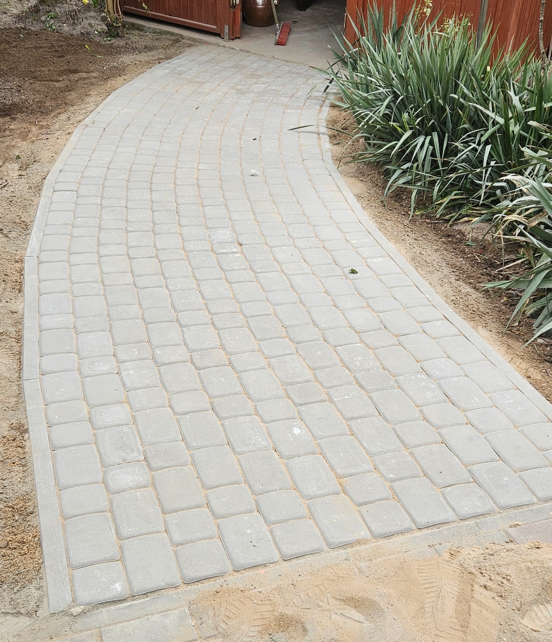 Zakrzywiona ścieżka z szarych kostek brukowych prowadząca do drewnianej bramy, obok rabata z zielonymi roślinami.