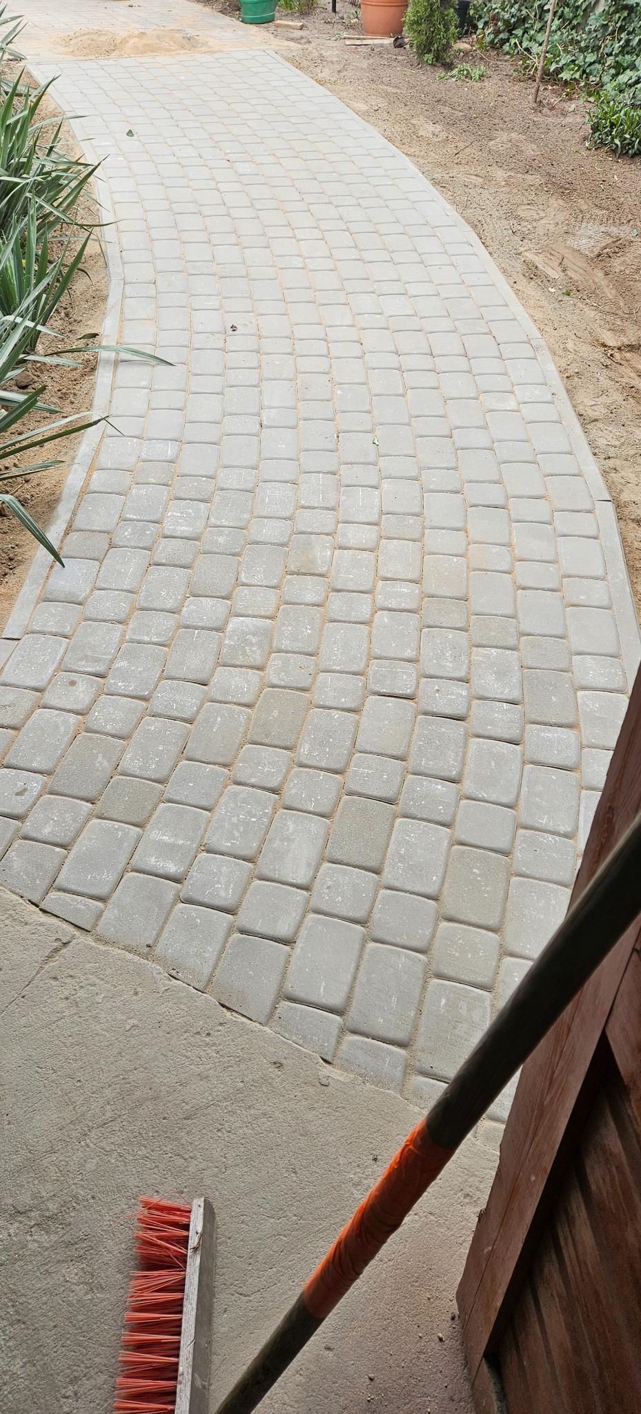 Ścieżka z szarej kostki brukowej ułożona łukiem, widoczny fragment szczotki z pomarańczową taśmą na trzonku oraz roślinność w tle.
