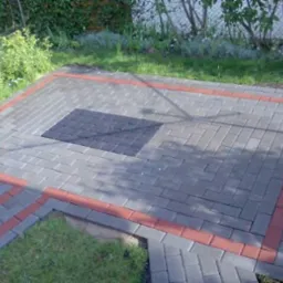 Wykończony taras z kostki brukowej w odcieniach szarości, z obramowaniem z czerwonej kostki. Centralny element z ciemniejszej kostki. Otoczenie: trawa i roślinność.