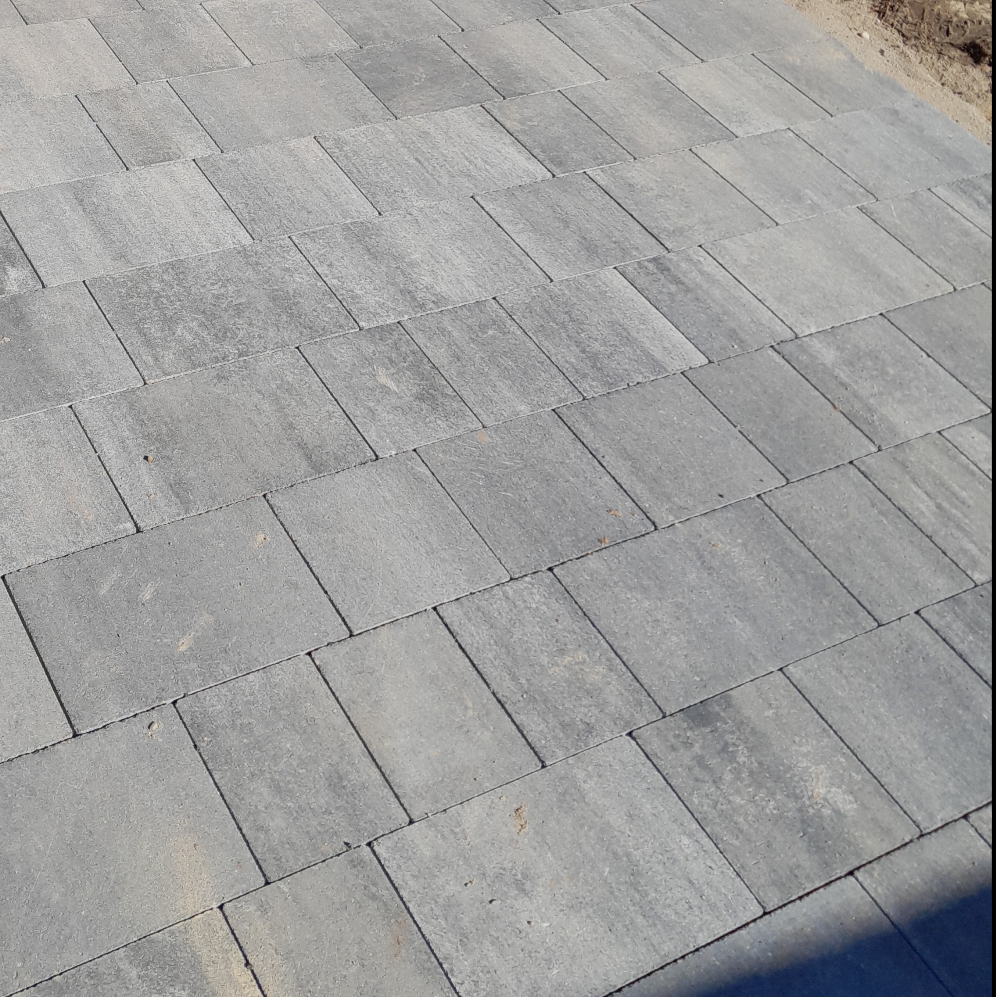 Ułożona szara kostka brukowa o prostokątnym kształcie, widoczna z góry pod kątem. Na obrzeżach widoczny piasek.