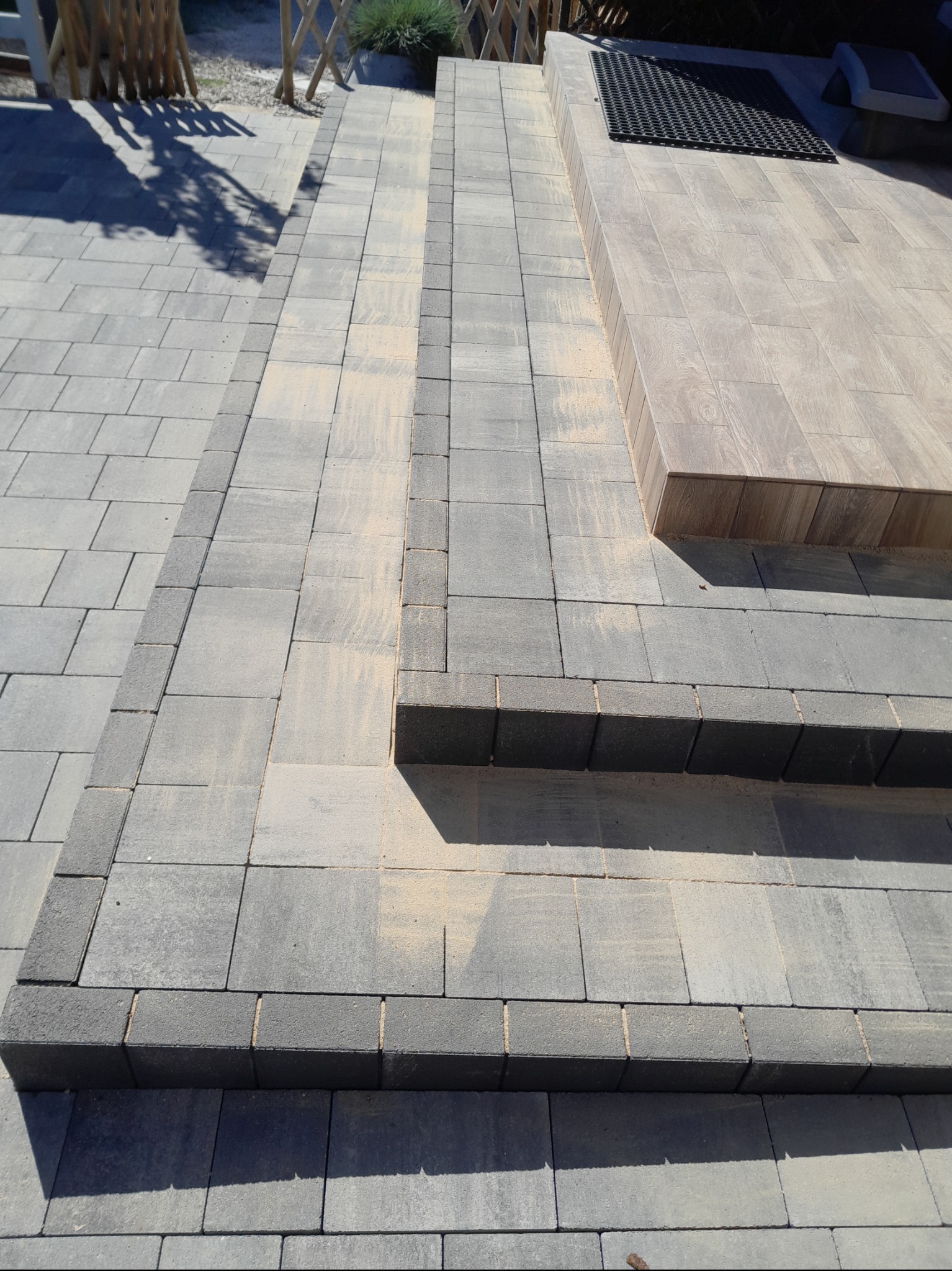 Schody z kostki brukowej w odcieniach szarości prowadzące do podestu z wycieraczką. Detal wykończenia z obrzeży. Widok z góry.