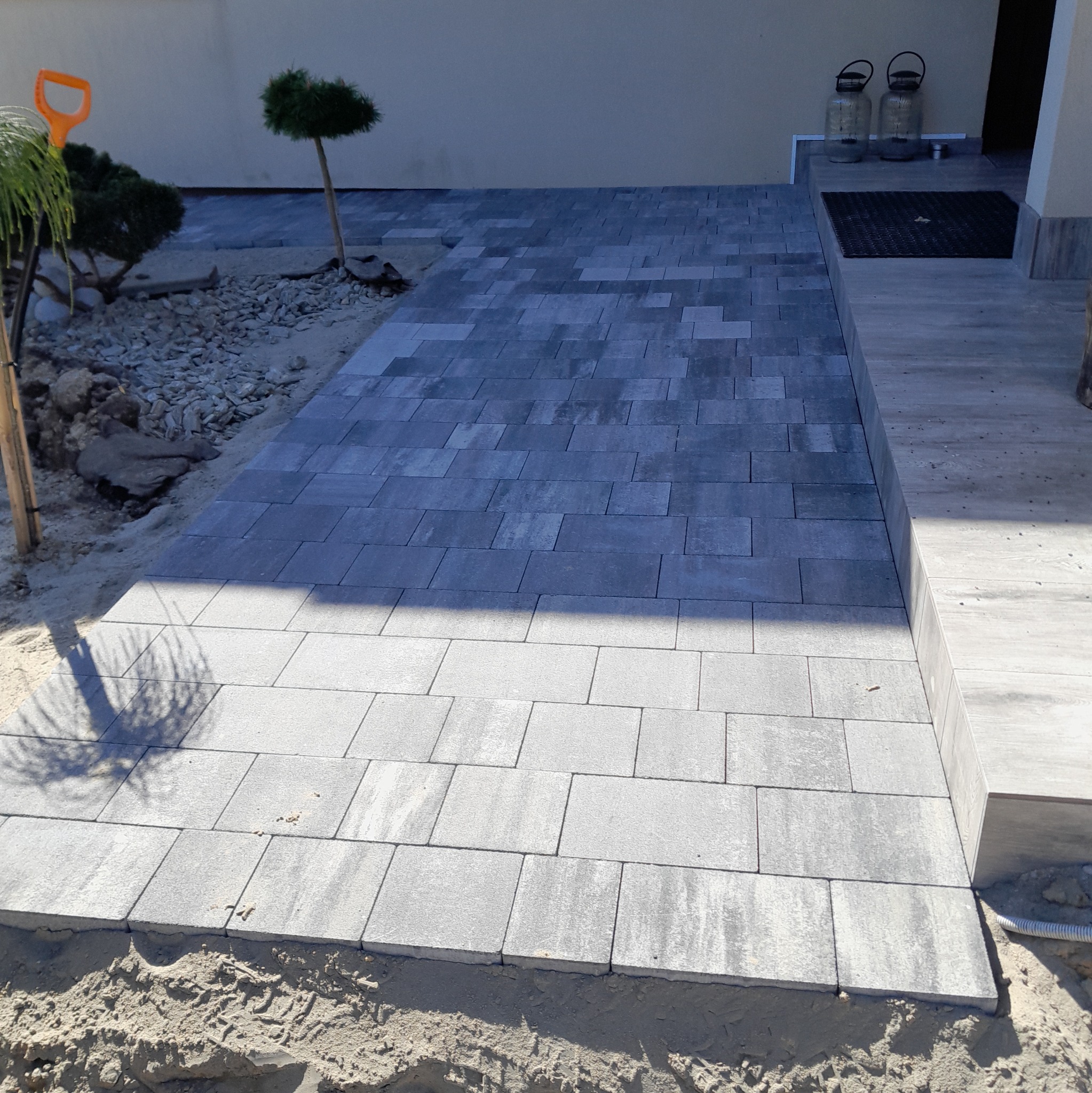 Świeżo ułożona, szara kostka brukowa prowadząca do wejścia domu, z widocznymi elementami krajobrazu i narzędziem ogrodniczym w tle.