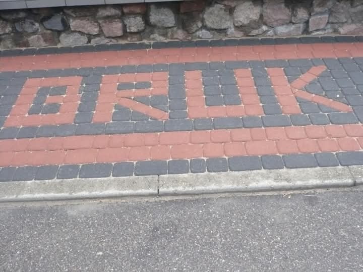 Ułożona kostka brukowa w kolorze czerwonym i szarym tworząca napis 'BRUK' na chodniku. Widoczne obrzeże chodnika i fragment kamiennego muru.