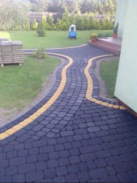 Ścieżka z ciemnej kostki brukowej z żółtymi obrzeżami prowadząca przez trawnik, w tle roślinność i zabawka dla dziecka. Widok na gotową realizację.