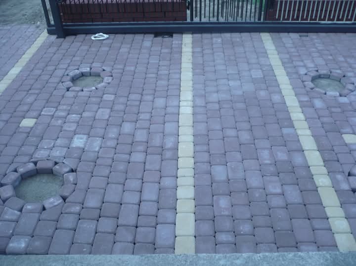 Ułożona kostka brukowa w odcieniach szarości z jasnymi pasami i okrągłymi otworami na rośliny, widziana z góry, przy bramie wjazdowej.