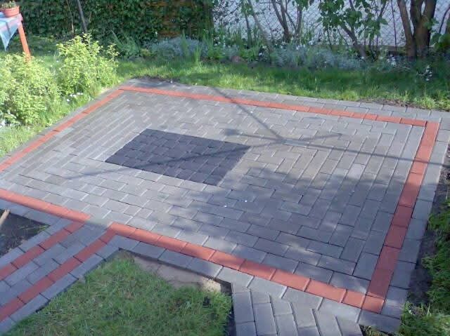 Wykończony taras z kostki brukowej w odcieniach szarości, z obramowaniem z czerwonej kostki. Centralny element z ciemniejszej kostki. Otoczenie: trawa i roślinność.