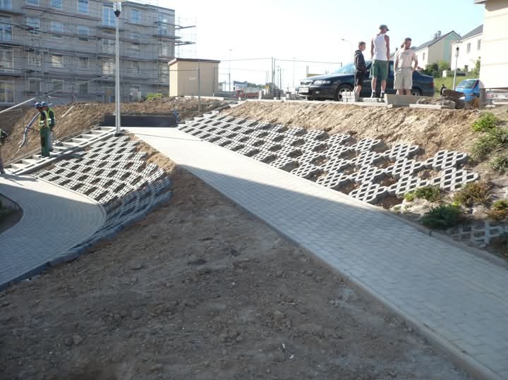 Szeroki kadr na ścieżkę z ażurowych płyt betonowych na zboczu, z pracownikami i budynkiem w tle, w słoneczny dzień.