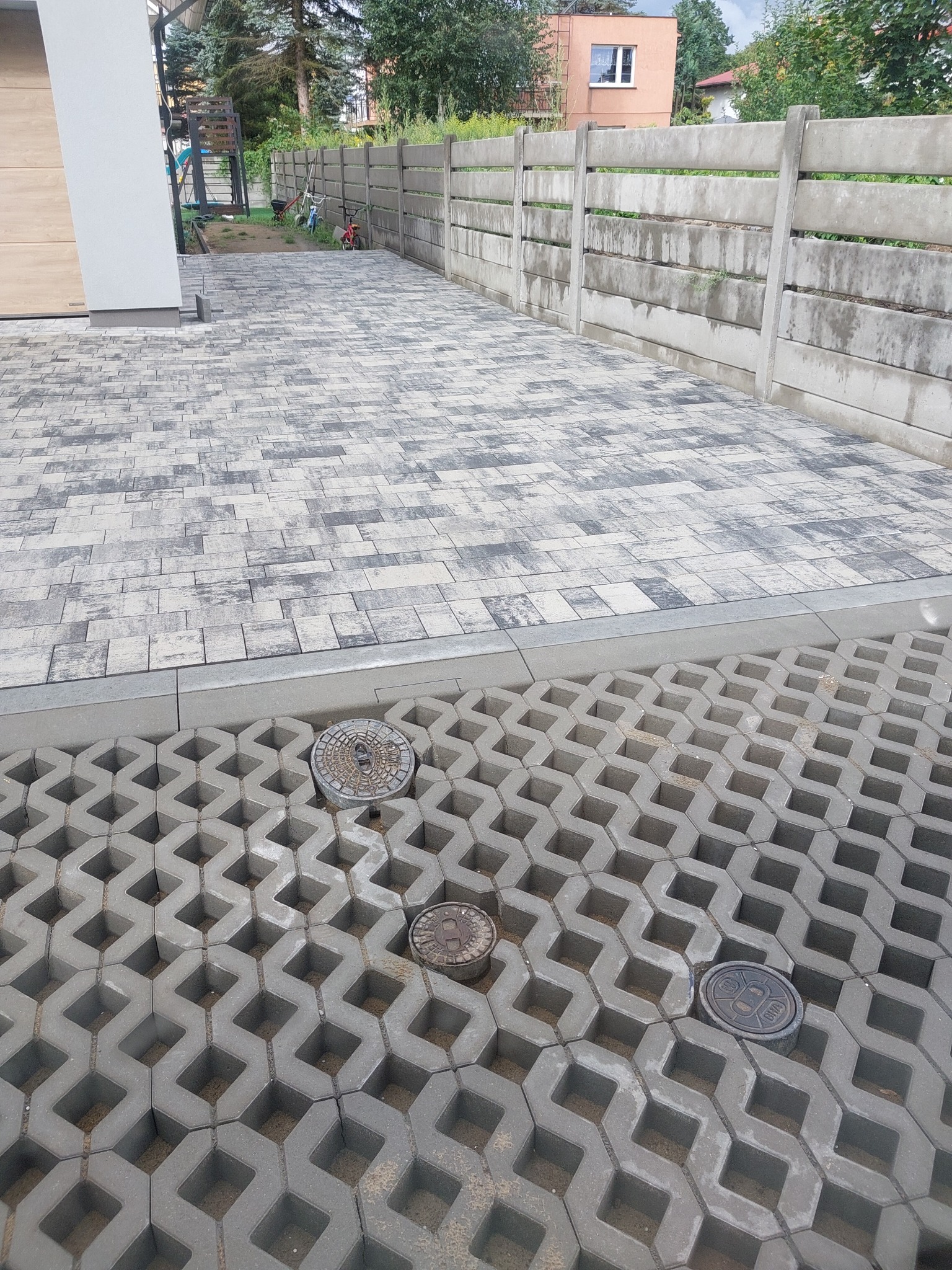 Ułożona szara kostka brukowa w dwóch wzorach, oddzielona obrzeżem. Widoczne studzienki kanalizacyjne i betonowy płot w tle.