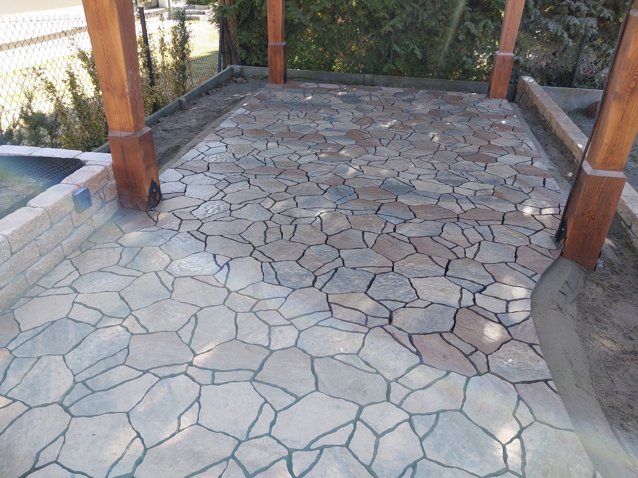 Ułożona nieregularna kostka kamienna tworząca taras z drewnianą pergolą. Widoczne detale łączeń i naturalna kolorystyka kamienia.