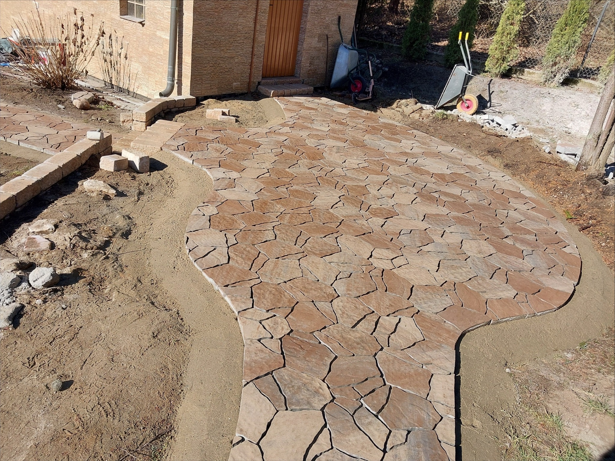 Kaskadowa ścieżka z nieregularnej, brązowej kostki, częściowo ułożona, z widocznymi narzędziami i elementami przygotowania podłoża. Widok z góry, słoneczny dzień.