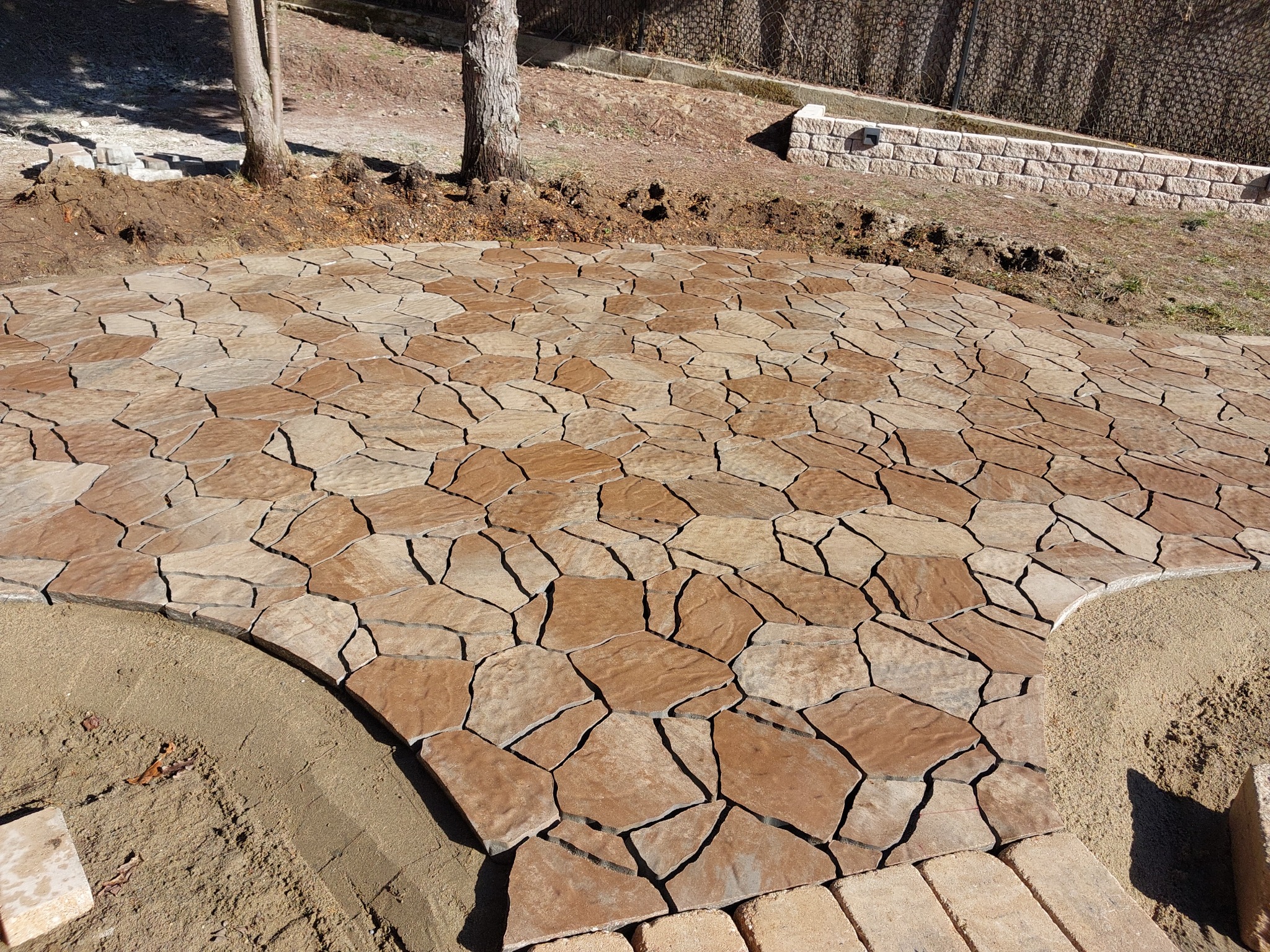 Ułożona, nieregularna kostka brukowa w odcieniach brązu, tworząca ścieżkę na posesji. Widoczne drzewa i przygotowane podłoże z piasku. Krawędzie wykończone kostką.