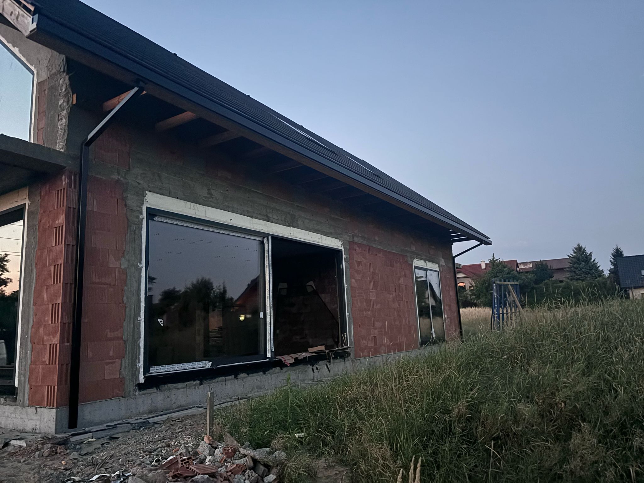 Surowy stan budynku z czerwonej cegły z zamontowanymi oknami, widoczne ślady zaprawy i gruz budowlany u podstawy, wysoka trawa w tle.