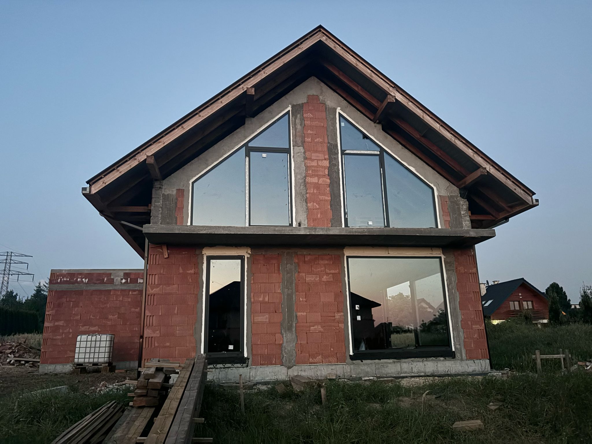 Surowy budynek w trakcie budowy z widocznymi oknami o nietypowym trójkątnym kształcie na poddaszu, cegły z betonu komórkowego, drewniane elementy konstrukcji dachu i niezagospodarowany teren wokół.