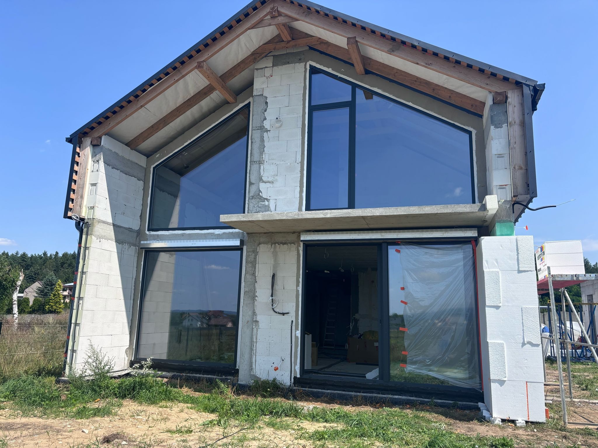 Dom w trakcie budowy z zamontowanymi oknami o dużych przeszkleniach i drzwiami tarasowymi, widoczne elementy konstrukcyjne dachu i ocieplenie ścian styropianem.