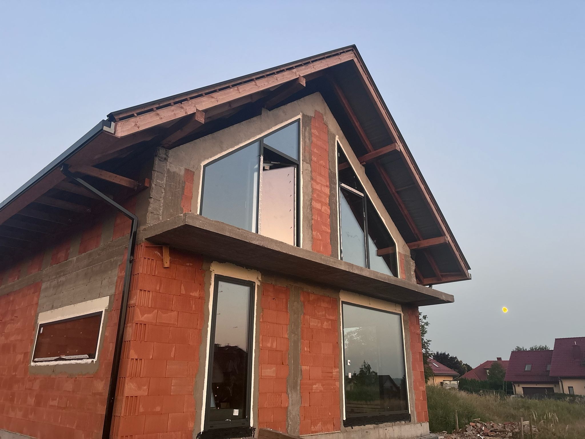 Surowy stan budynku z czerwonej cegły z zamontowanymi oknami o czarnych ramach, widoczne elementy drewnianej konstrukcji dachu i betonowy gzyms, w oknach odbija się zachodzące słońce.