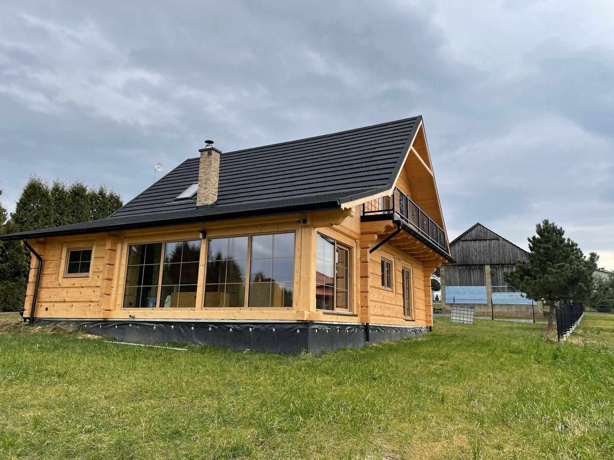 Nowoczesny dom z bali z dużymi oknami i balkonem, wykończony czarną dachówką, widok z zewnątrz na tle pochmurnego nieba.