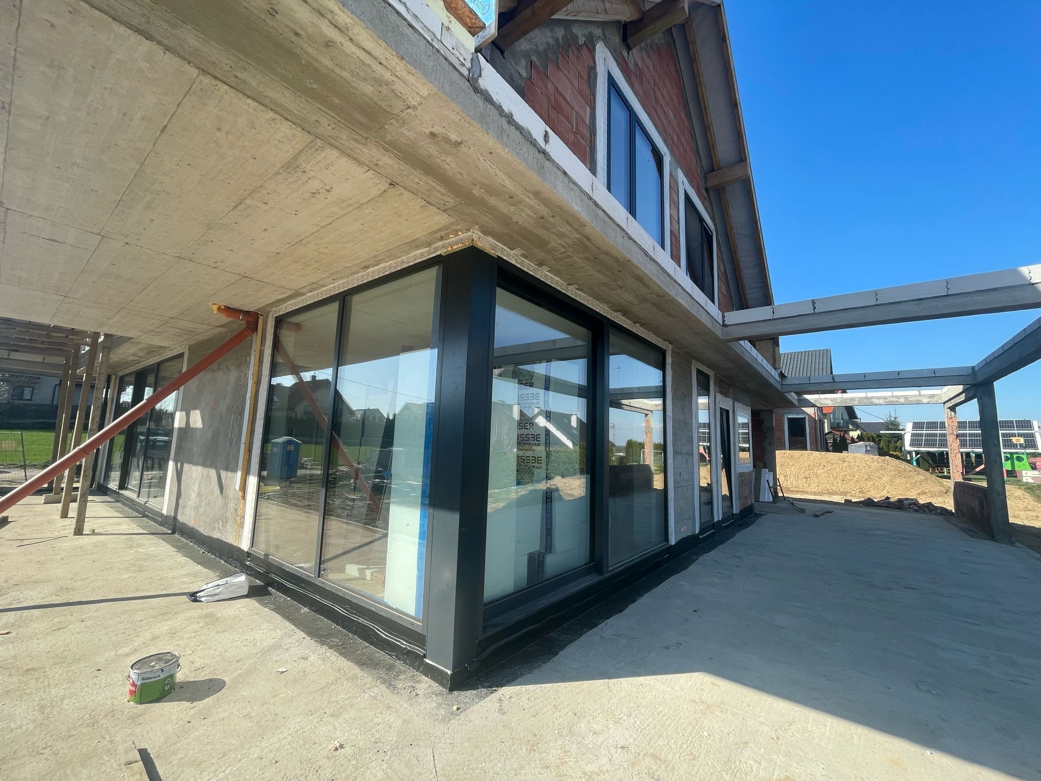 Surowy stan budynku z zamontowanymi dużymi oknami z czarnymi ramami, widoczne elementy konstrukcyjne i zaplecze budowy.