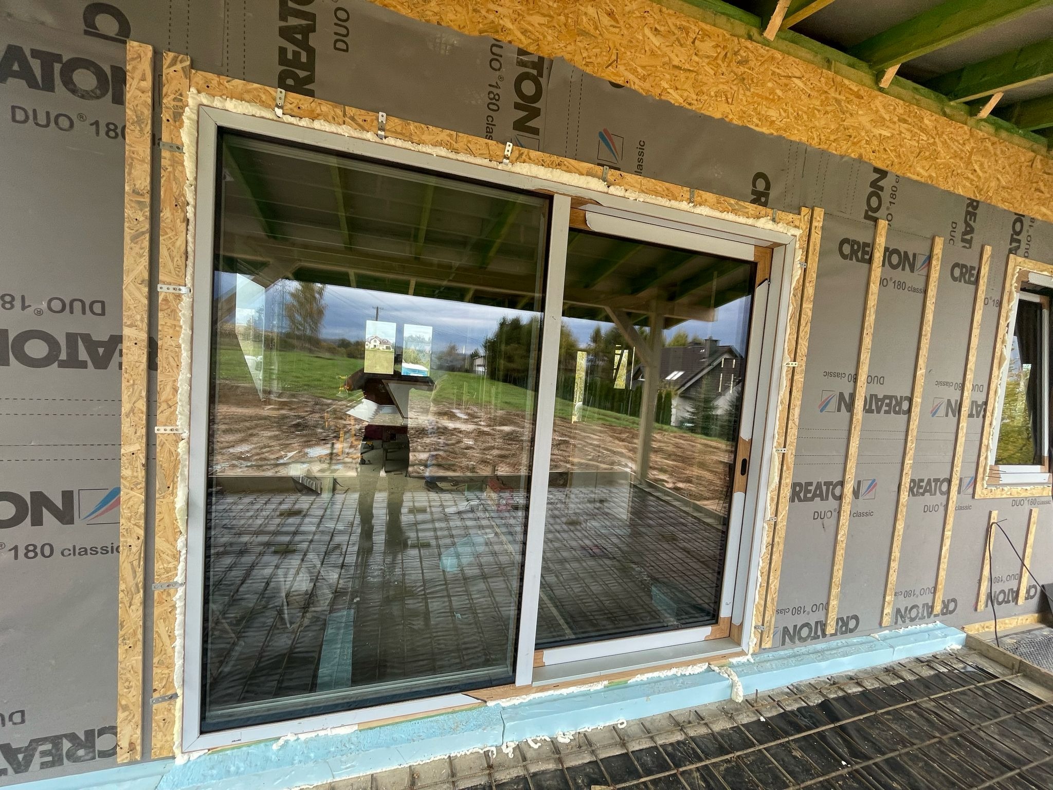 Wstawione okno przesuwne z widokiem na zewnątrz, osadzone w surowej ramie z pianką montażową i folią izolacyjną, widoczna konstrukcja pod wylewkę betonową.