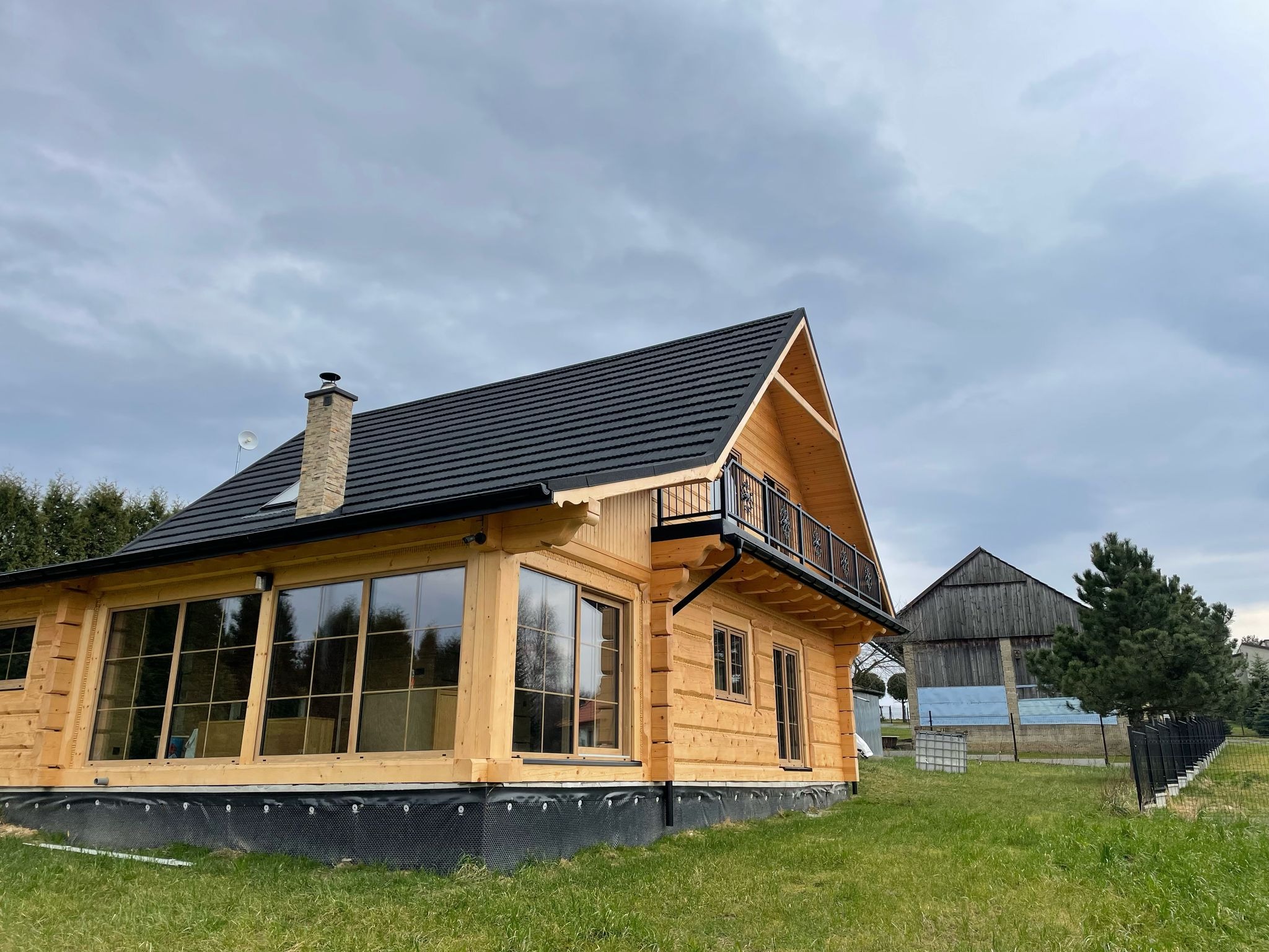 Nowoczesny dom z bali z czarną dachówką i małym balkonem, widok z zewnątrz na tle pochmurnego nieba i starej stodoły.