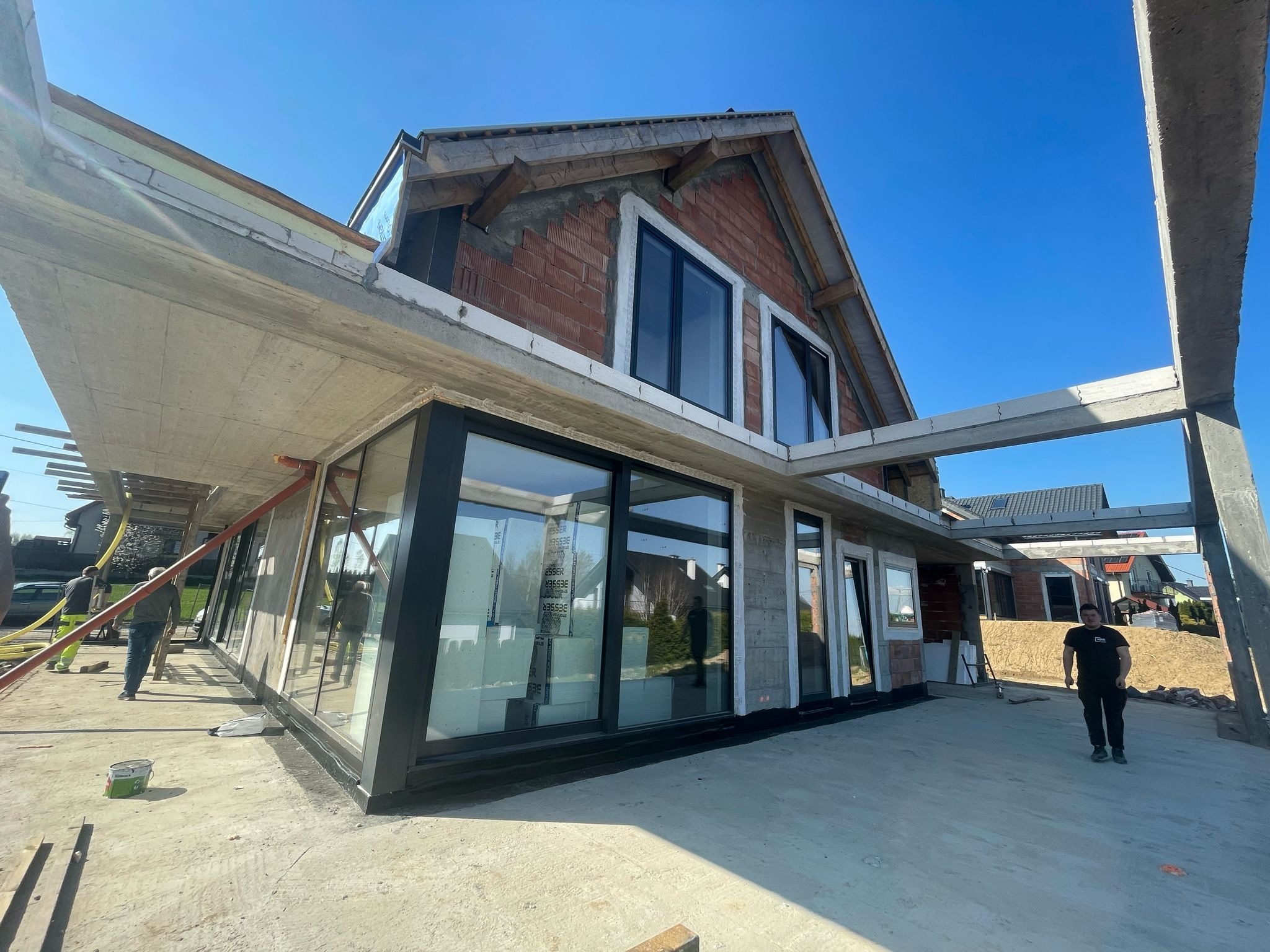 Surowy stan budynku z widocznymi nowymi oknami z czarnymi ramami, ściany z czerwonej cegły, betonowe elementy konstrukcyjne i pracownicy na zewnątrz.