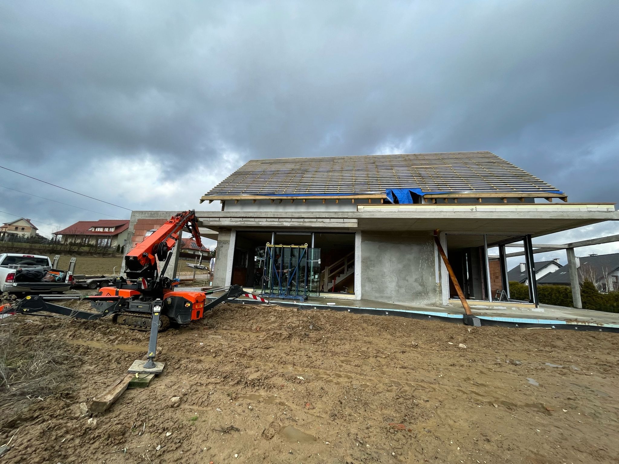 Budowa domu jednorodzinnego w stanie surowym otwartym; widoczny dźwig budowlany na błotnistym terenie, przygotowywane otwory okienne z zamontowanymi ramami, częściowo deskowany dach pod przykryciem...
