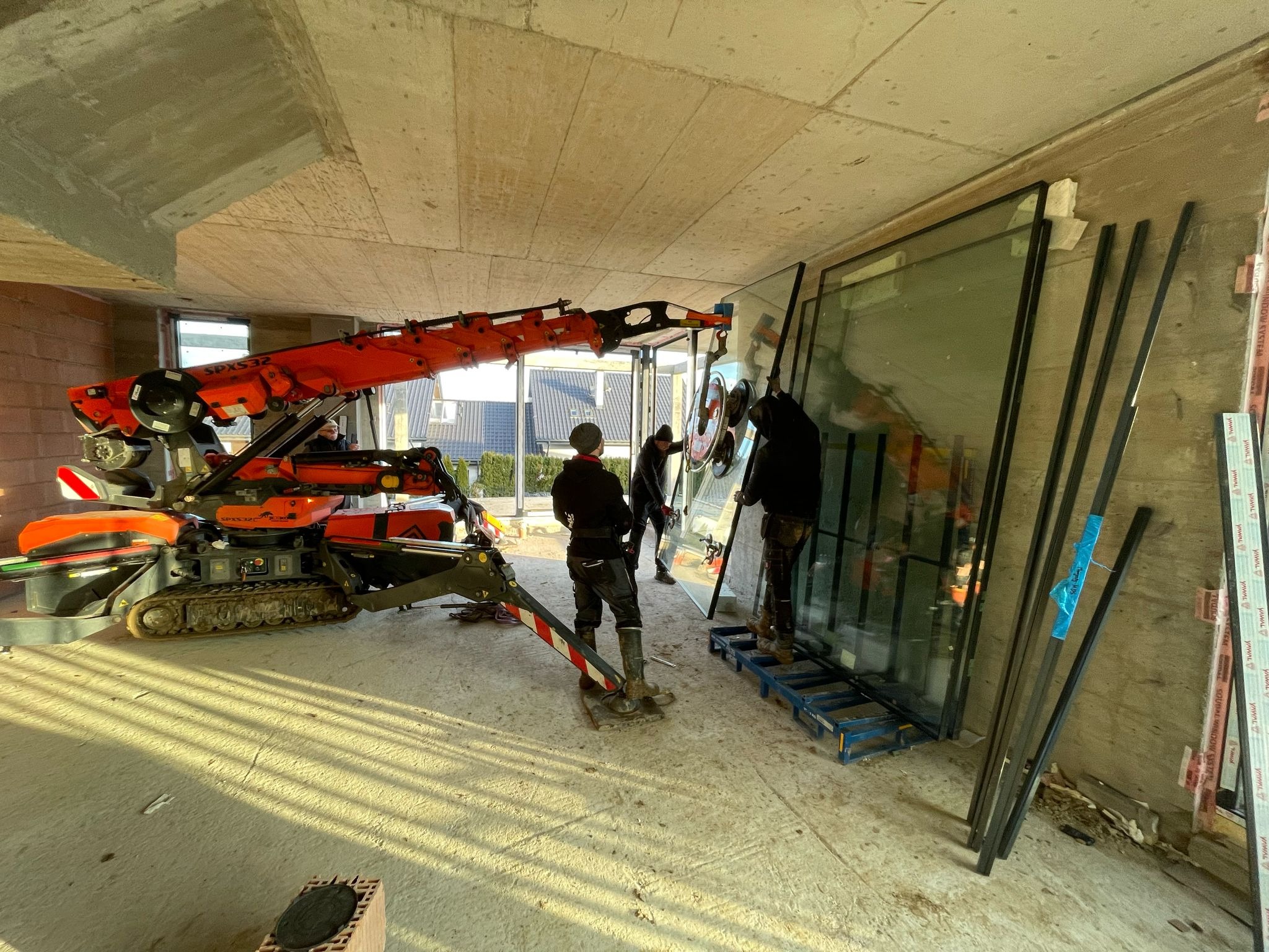 Montaż dużych okien z czarnymi ramami za pomocą specjalistycznego dźwigu SPX532 w surowym stanie budynku; widoczni trzej monterzy, jeden z nich używa przyssawek do szkła.