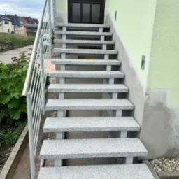 Zewnętrzne schody wejściowe wyłożone szarym granitem z metalową balustradą, prowadzące do ciemnych drzwi w jasnozielonej elewacji budynku.