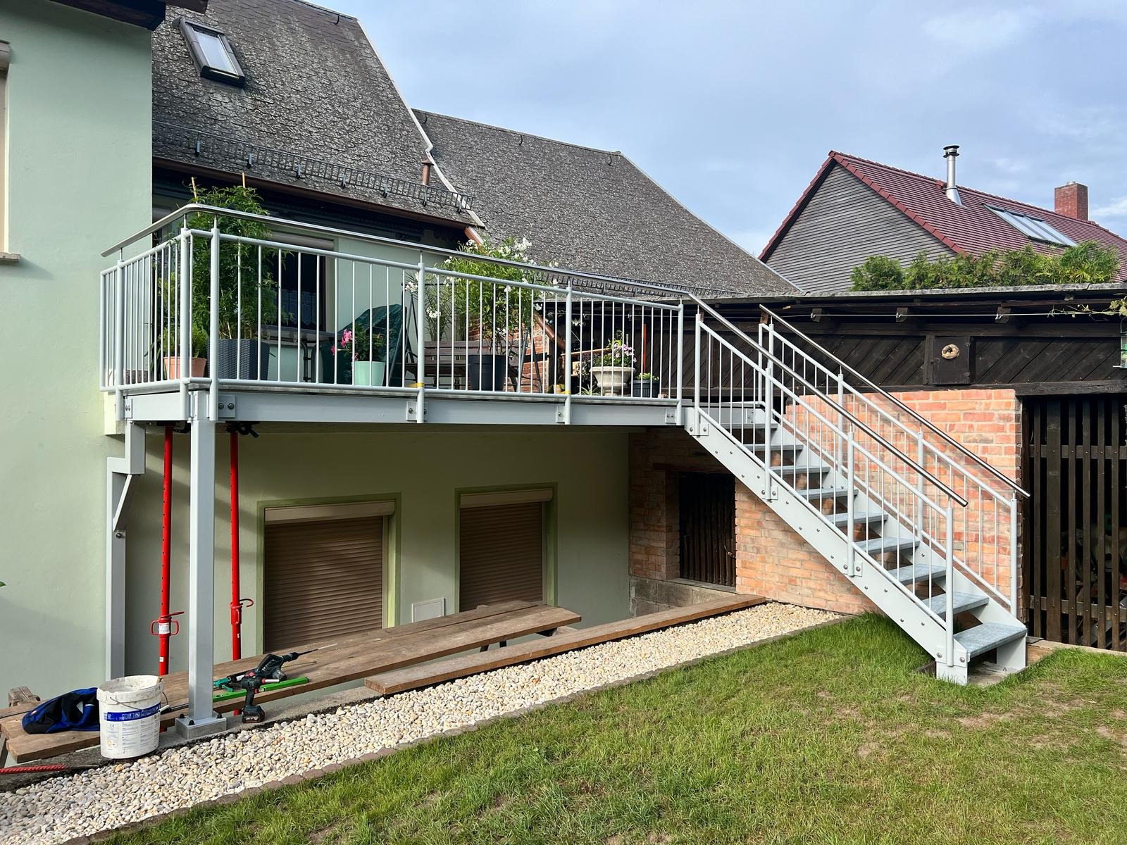 Zewnętrzny metalowy balkon z białymi barierkami i schodami, wsparty na czerwonych podporach, z widocznymi narzędziami i materiałami budowlanymi u podstawy, sugerującymi trwający montaż.