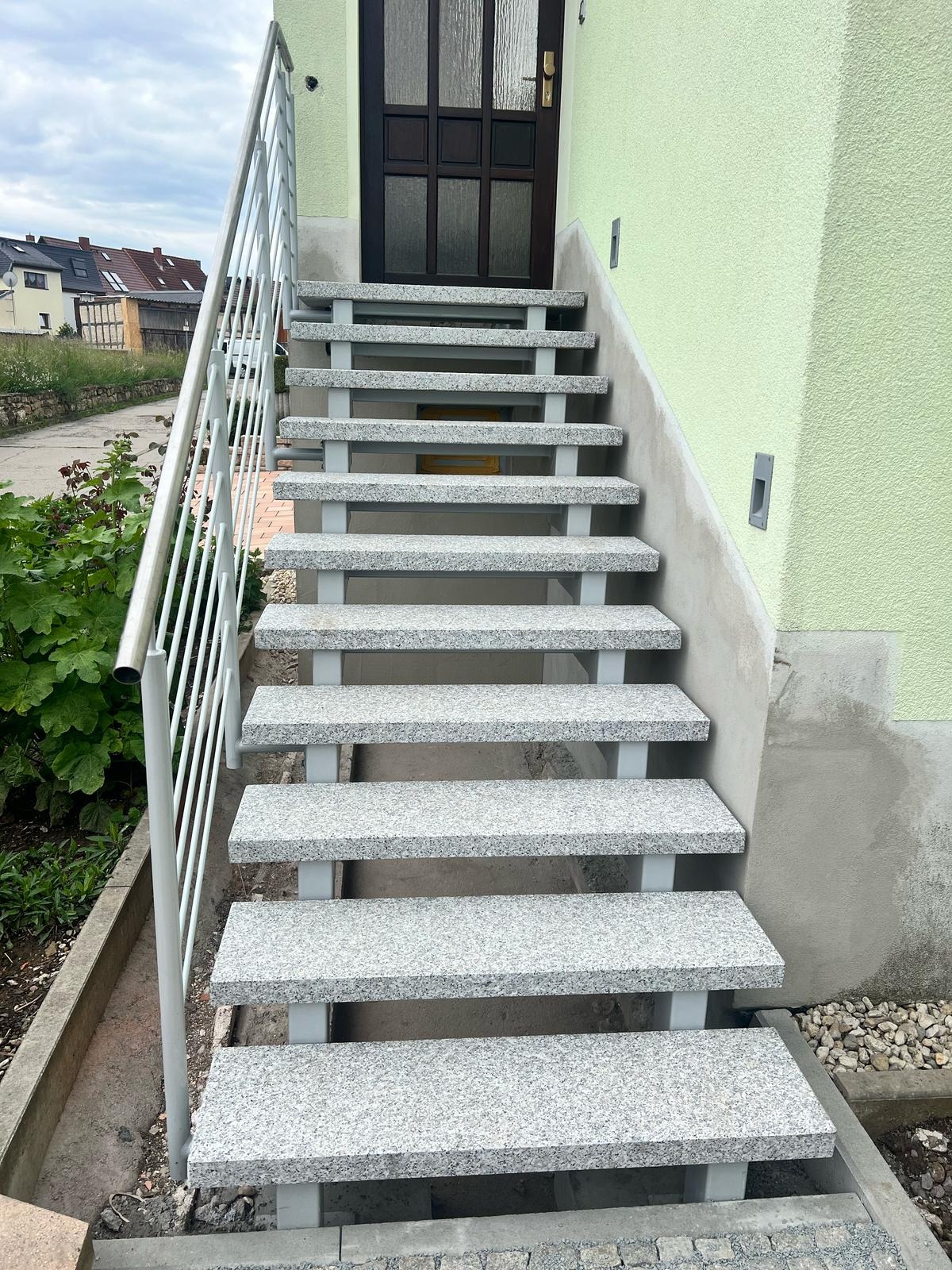 Zewnętrzne schody wejściowe wyłożone szarym granitem z metalową balustradą, prowadzące do ciemnych drzwi w jasnozielonej elewacji budynku.