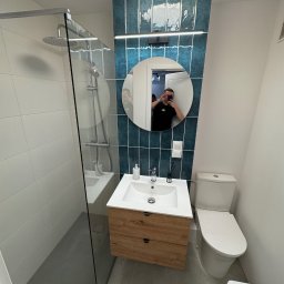 Veltrix - Nowoczesna łazienka z kabiną prysznicową typu walk-in, umywalką z szafką z naturalnego drewna, okrągłym lustrem z odbiciem fotografa i pionowymi, błyszczącymi płytkami w odcieniach morskiej zieleni.