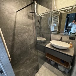 Veltrix - Nowoczesna łazienka z kabiną prysznicową typu walk-in, szare płytki imitujące kamień na ścianach i podłodze, owalna umywalka nablatowa na szafce z otwartą półką, okrągłe lustro z podświetleniem LED.