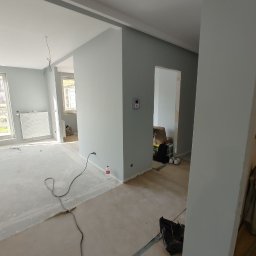 Veltrix - Wnętrze w trakcie prac wykończeniowych: świeżo pomalowane ściany w odcieniach szarości, widoczne kable elektryczne, zabezpieczona podłoga, butelka wody i torba z narzędziami w tle.