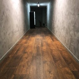 Veltrix - Długi korytarz z nową, ciemną, drewnianą podłogą i szarymi ścianami o fakturze betonu, zakończony automatem z napojami i gaśnicą w tle.