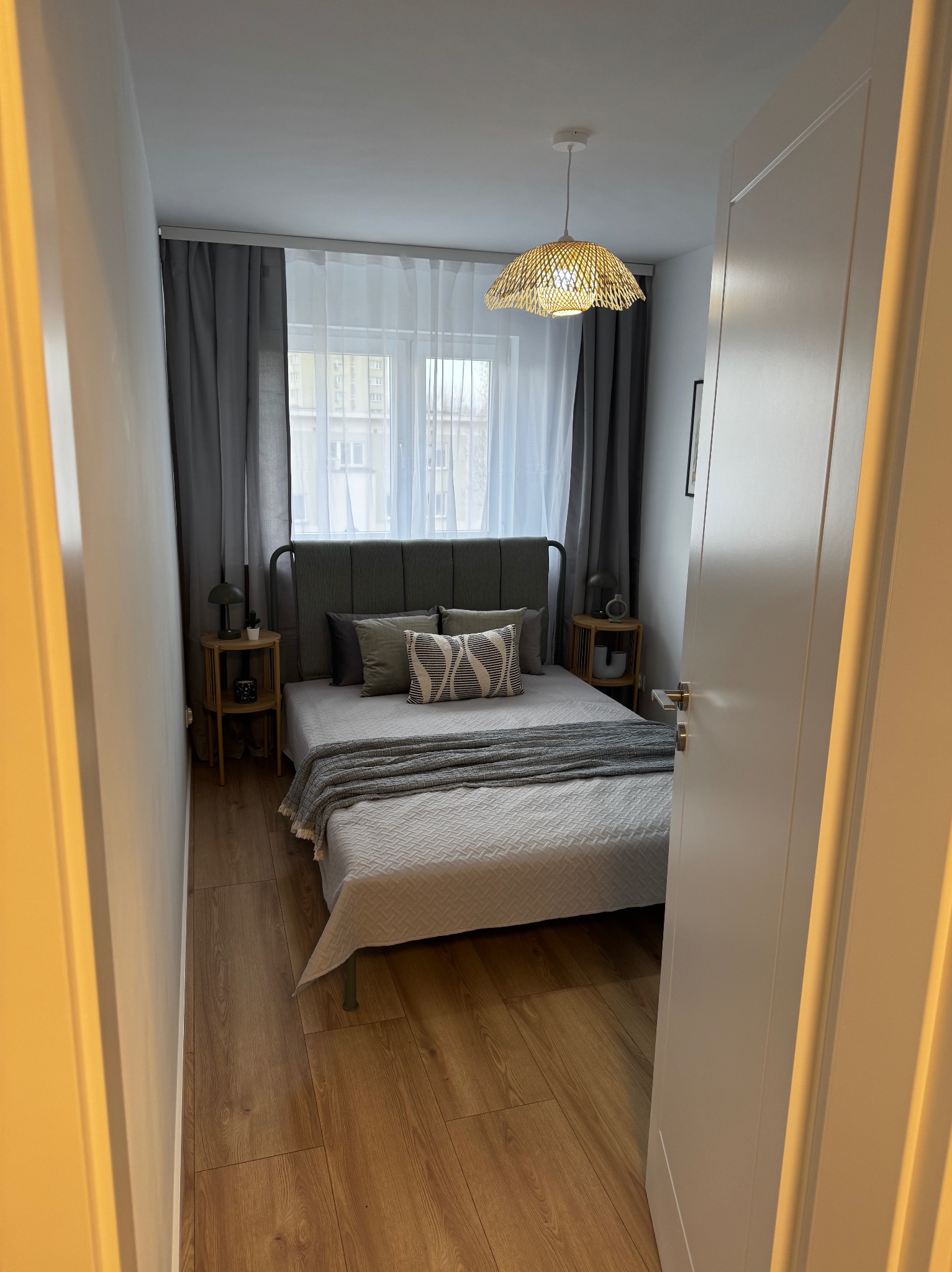 Ustawna sypialnia z łóżkiem w szarościach, drewnianą podłogą i plecioną lampą, widok zza uchylonych, białych drzwi.