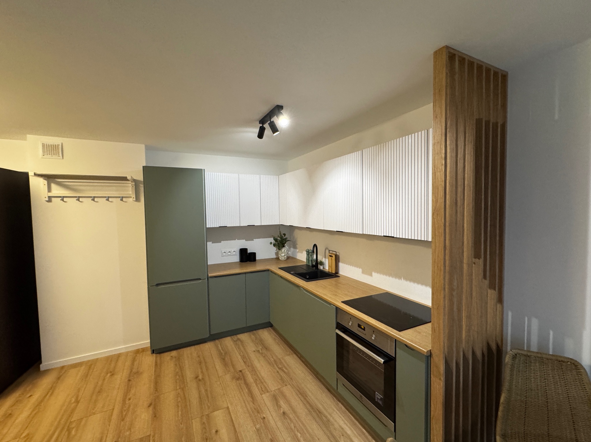 Nowoczesna kuchnia z oliwkowymi szafkami dolnymi i białymi górnymi z pionowymi frezami, drewnianym blatem i podłogą, czarną armaturą oraz drewnianą ażurową ścianką działową.