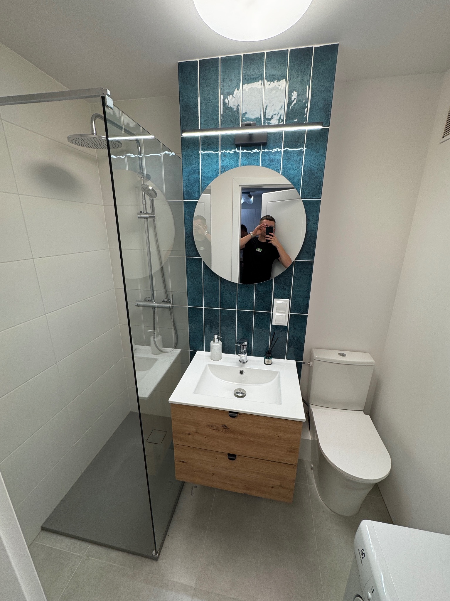 Nowoczesna łazienka z kabiną prysznicową typu walk-in, umywalką z szafką z naturalnego drewna, okrągłym lustrem z odbiciem fotografa i pionowymi, błyszczącymi płytkami w odcieniach morskiej zieleni.