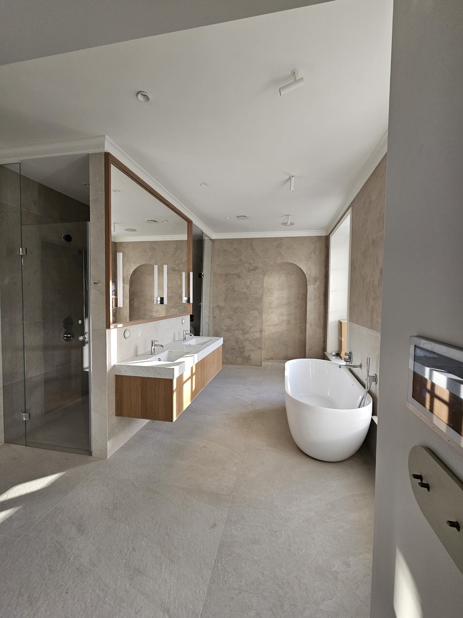 Nowoczesna łazienka z wolnostojącą wanną, podwójną umywalką z drewnianą szafką i przeszklonym prysznicem, wykończona w beżowych tonacjach z dekoracyjnymi wnękami w ścianie.