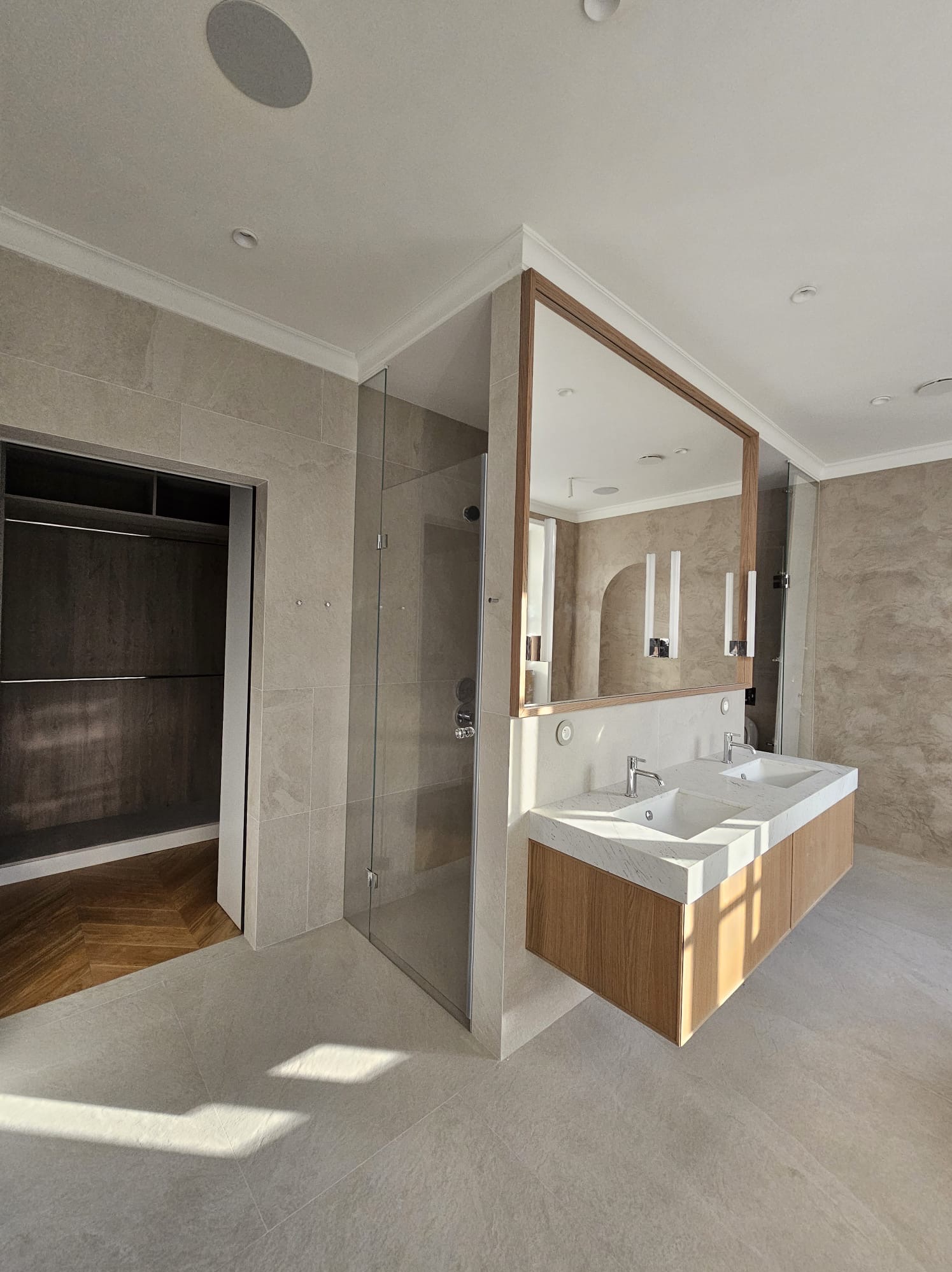 Jasna łazienka z dwoma umywalkami na blacie z imitacji marmuru, kabina prysznicowa ze szklanymi drzwiami i wejście do garderoby z parkietem w jodełkę.