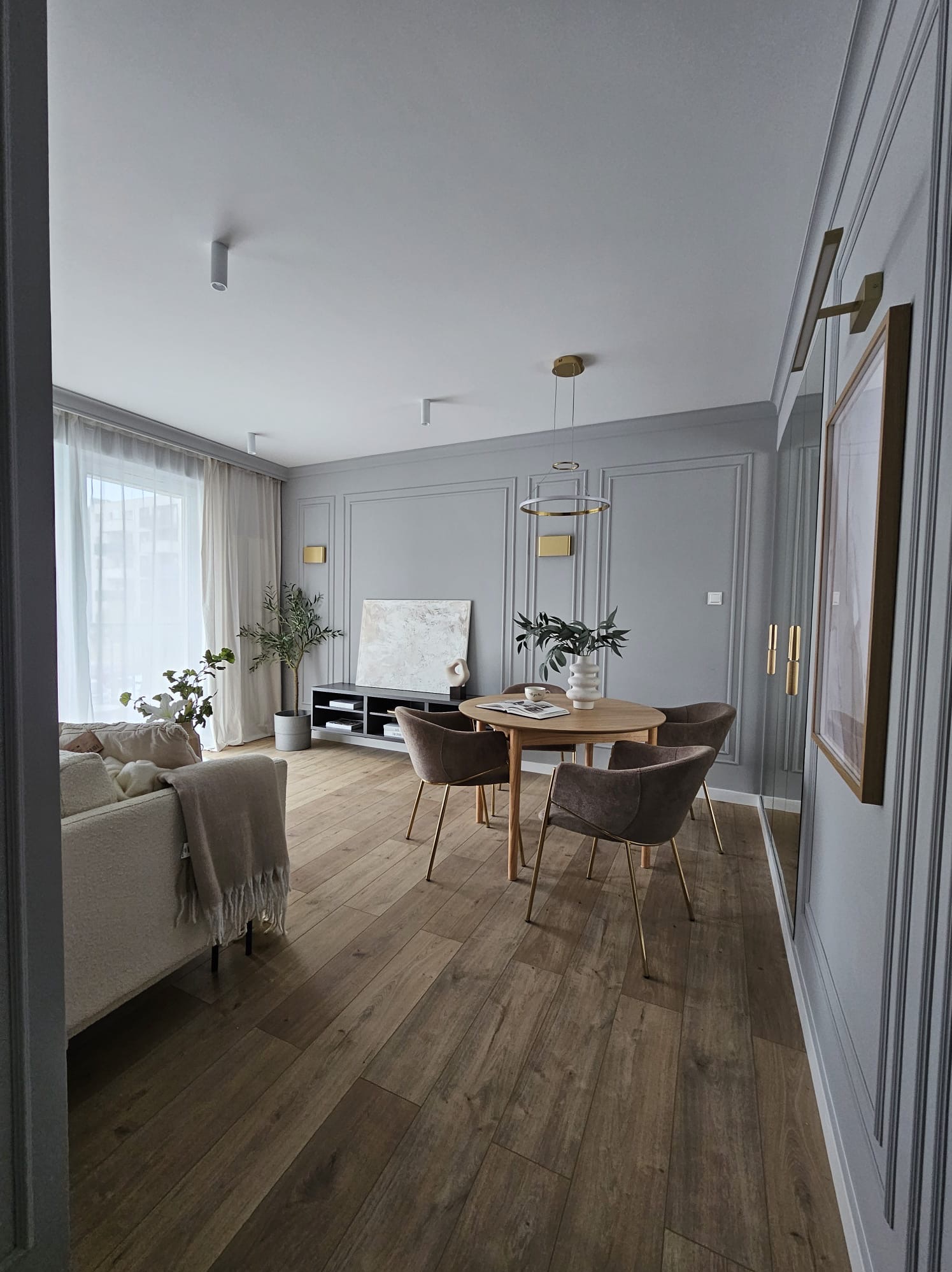 Jasne, nowocześnie urządzone wnętrze salonu z drewnianą podłogą, szarymi ścianami z dekoracyjnymi listwami, okrągłym drewnianym stołem z czterema krzesłami i beżową sofą z narzutą.