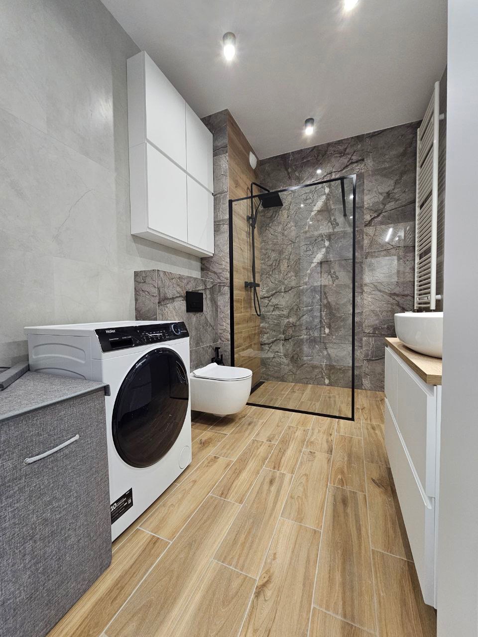 Nowoczesna łazienka z drewnopodobnymi płytkami, kabiną prysznicową typu walk-in z czarną ramą, wiszącą toaletą, pralką i białymi szafkami.