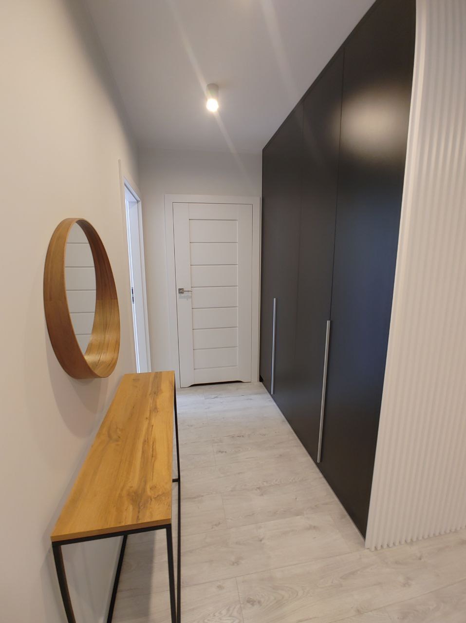 Nowoczesny przedpokój z drewnianym okrągłym lustrem, konsolą na metalowych nogach, białą stolarką drzwiową i antracytową szafą w zabudowie.