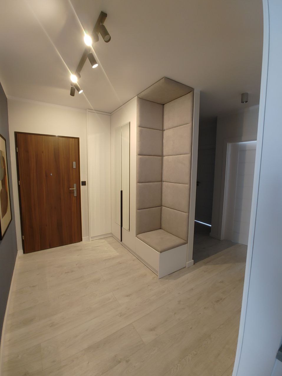 Nowoczesny przedpokój z drewnianymi drzwiami, białymi szafami w zabudowie i tapicerowanym siedziskiem w rogu, oświetlony punktowymi lampami sufitowymi.