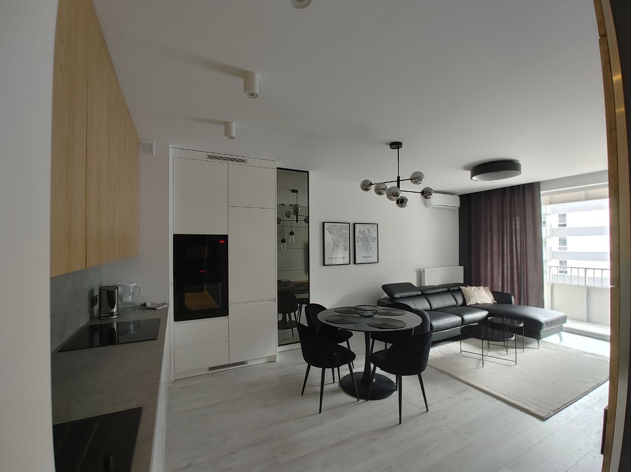 Nowoczesne wnętrze mieszkania z jasną podłogą, czarną sofą narożną, okrągłym stołem z krzesłami i zabudową kuchenną z białymi frontami i czarnym piekarnikiem.