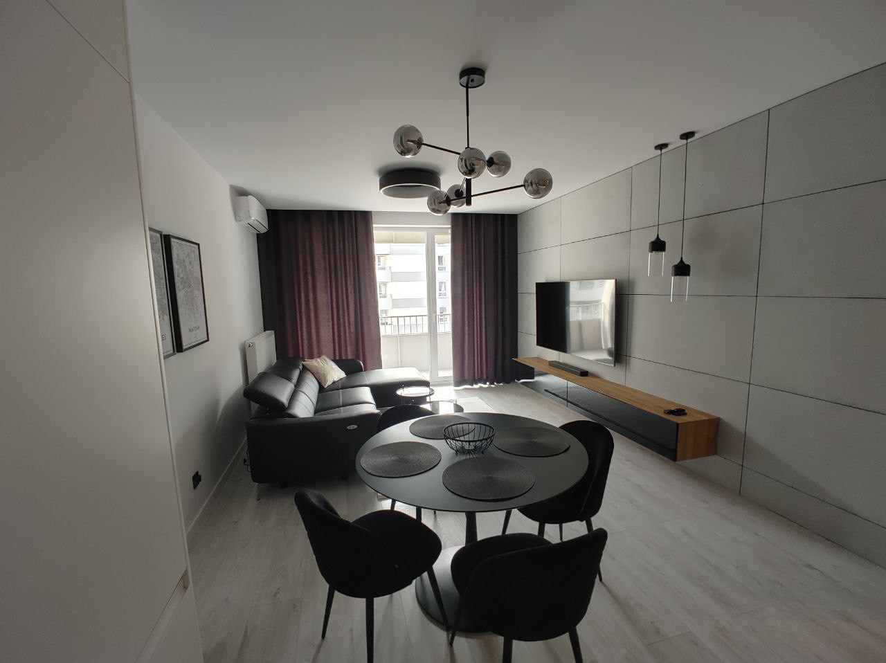 Nowoczesny salon z szarą ścianą z płyt, czarną sofą narożną, okrągłym stołem z czarnymi krzesłami i designerską lampą wiszącą.