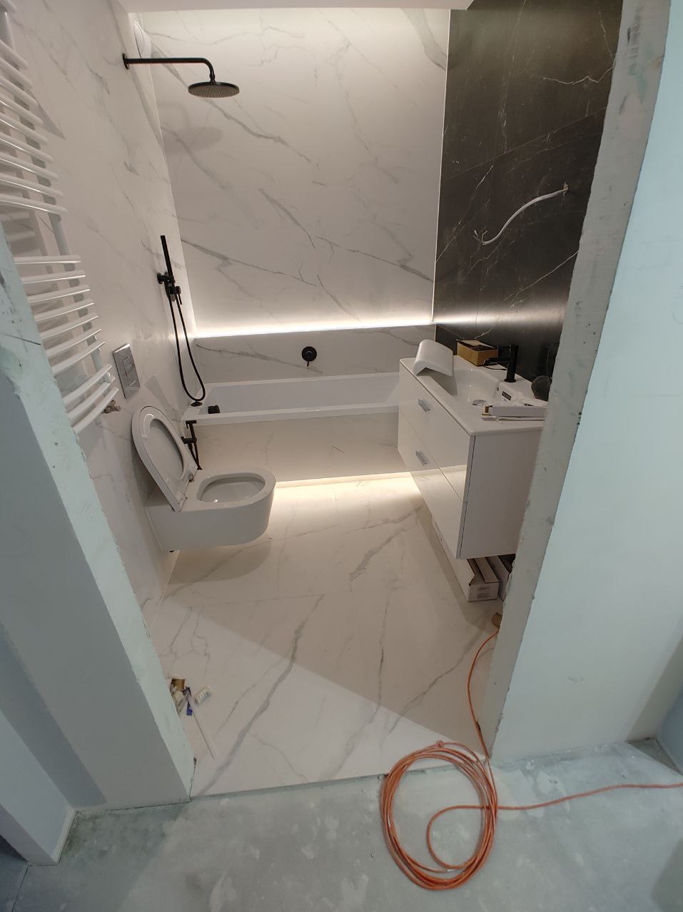 Nowoczesna łazienka w trakcie wykańczania: białe marmurowe płytki, czarna armatura, podświetlana wanna i szafka umywalkowa, widoczne niedokończone elementy wykończenia i zwój pomarańczowego kabla.